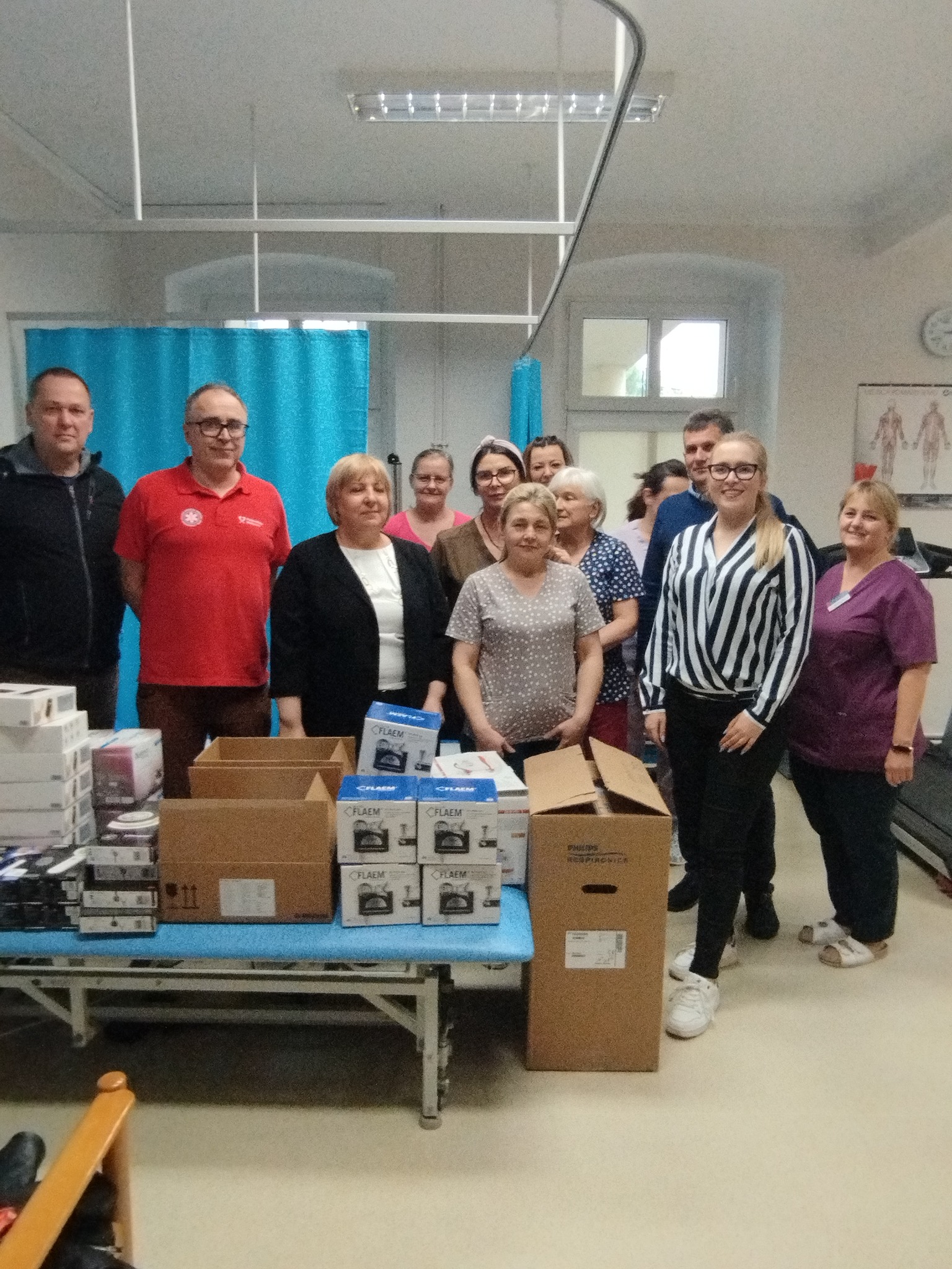 Grupa osób stojących za stołem z kartonami i opakowaniami sprzętu medycznego w pomieszczeniu szpitalnym.