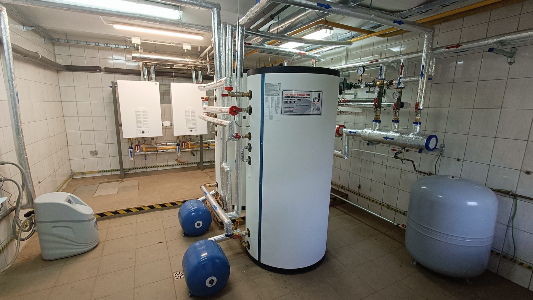 Pomieszczenie techniczne z instalacją grzewczą, zawierające zbiornik buforowy, dwa białe kotły oraz orurowanie